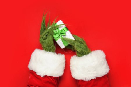 Téléchargez les photos : Mains poilues vertes de créature en costume de Père Noël avec boîte cadeau sur fond rouge - en image libre de droit