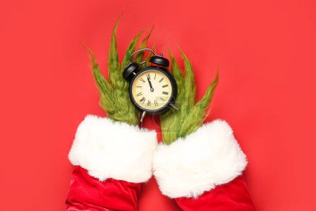 Téléchargez les photos : Mains poilues vertes de créature en costume de Père Noël avec réveil sur fond rouge - en image libre de droit