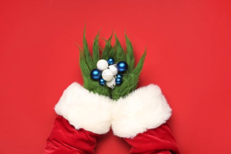 Photo for Hairy hands of green creature in Santa costume with Christmas balls on red background - Royalty Free Image