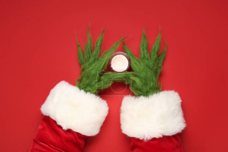 Téléchargez les photos : Mains poilues vertes de créature en costume de Père Noël avec bougie sur fond rouge - en image libre de droit
