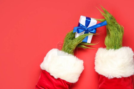 Téléchargez les photos : Mains poilues vertes de créature en costume de Père Noël avec boîte cadeau sur fond rouge - en image libre de droit