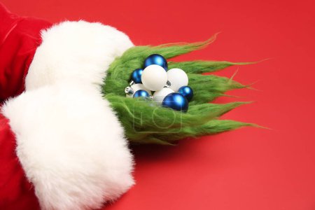 Foto de Peludas manos de criatura verde en traje de Santa con bolas de Navidad sobre fondo rojo - Imagen libre de derechos