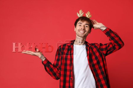 Téléchargez les photos : Jeune homme tatoué dans des cornes de renne avec salutation de Noël sur fond rouge - en image libre de droit