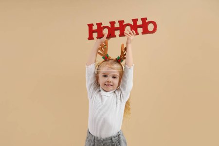 Téléchargez les photos : Petite fille mignonne en cornes de rennes avec salutation de Noël sur fond beige - en image libre de droit