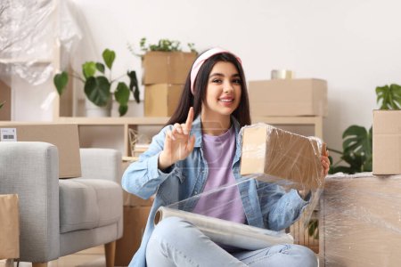 Téléchargez les photos : Jeune femme enveloppant boîte en carton avec film étirable à la maison le jour du déménagement - en image libre de droit