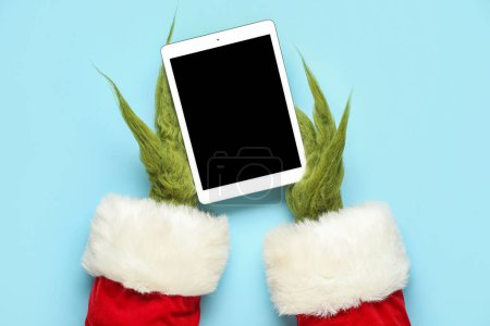 Téléchargez les photos : Mains poilues vertes de créature en costume de Père Noël avec tablette sur fond bleu - en image libre de droit