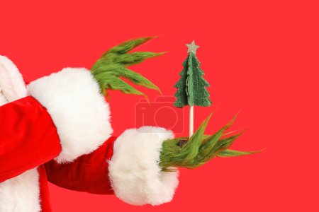 Photo for Green hairy hands of creature in Santa costume with decorative fir tree on red background - Royalty Free Image