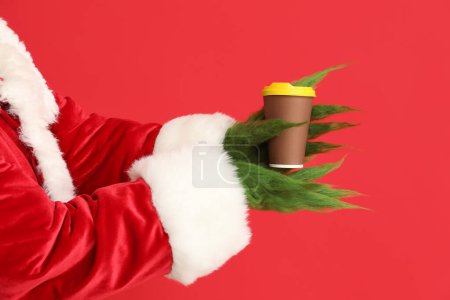 Téléchargez les photos : Créature poilue verte en costume de Père Noël avec tasse de café sur fond rouge - en image libre de droit