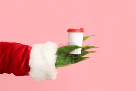 Téléchargez les photos : Main poilue verte de créature en costume de Père Noël avec café sur fond rose - en image libre de droit