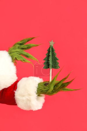 Téléchargez les photos : Mains poilues vertes de créature en costume de Père Noël avec sapin décoratif sur fond rouge - en image libre de droit