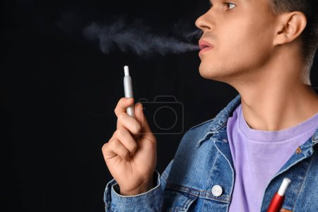 Téléchargez les photos : Jeune homme fumant cigarette électronique sur fond sombre - en image libre de droit