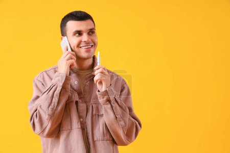 Téléchargez les photos : Jeune homme avec cigarette électronique parlant par téléphone sur fond jaune - en image libre de droit
