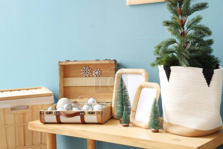 Photo for Wooden bench with blank frames, mini Christmas trees and baubles in suitcase near blue wall, closeup - Royalty Free Image