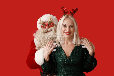 Téléchargez les photos : Père Noël et belle femme mature dans les cornes de cerf de Noël sur fond rouge - en image libre de droit