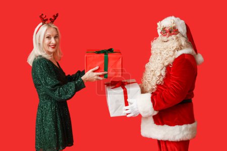 Téléchargez les photos : Père Noël et belle femme mature dans des cornes de cerf de Noël avec des boîtes-cadeaux sur fond rouge - en image libre de droit