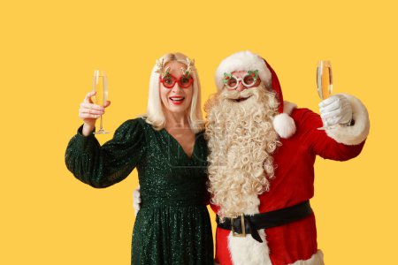 Téléchargez les photos : Père Noël et belle femme mûre dans des verres de Noël avec champagne sur fond jaune - en image libre de droit