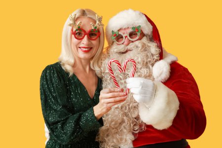 Téléchargez les photos : Père Noël et belle femme mature dans des lunettes de Noël avec des cannes à bonbons sur fond jaune - en image libre de droit