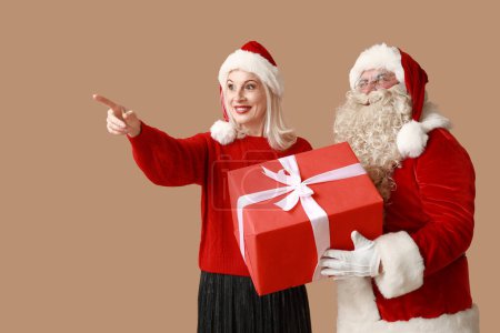 Téléchargez les photos : Père Noël et belle femme mature dans des chapeaux de Noël avec boîte cadeau pointant quelque chose sur fond brun - en image libre de droit