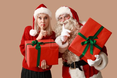 Téléchargez les photos : Père Noël et belle femme mature dans des chapeaux de Noël avec des boîtes-cadeaux montrant geste silencieux sur fond brun - en image libre de droit
