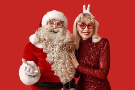 Téléchargez les photos : Père Noël et belle femme mûre dans des lunettes de Noël montrant geste de victoire sur fond rouge - en image libre de droit
