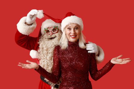Téléchargez les photos : Père Noël avec belle femme mature dans des lunettes de Noël et chapeaux sur fond rouge - en image libre de droit