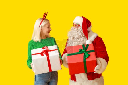 Téléchargez les photos : Père Noël et belle femme mature dans des cornes de cerf de Noël avec des boîtes-cadeaux sur fond jaune - en image libre de droit