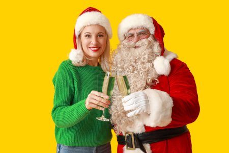 Téléchargez les photos : Père Noël et belle femme mature dans des chapeaux de Noël avec des verres de champagne sur fond jaune - en image libre de droit