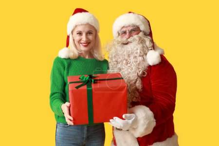 Téléchargez les photos : Père Noël et belle femme mature dans des chapeaux de Noël avec boîte cadeau sur fond jaune - en image libre de droit