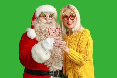 Téléchargez les photos : Père Noël et belle femme mature dans des lunettes de Noël avec des cannes à bonbons sur fond vert - en image libre de droit