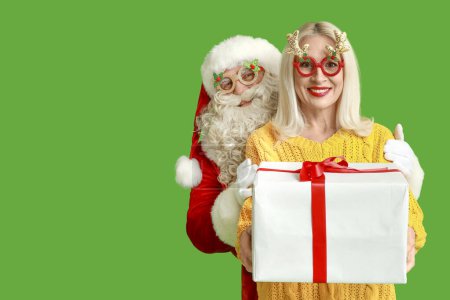 Téléchargez les photos : Père Noël et belle femme mature dans des lunettes de Noël avec boîte cadeau sur fond vert - en image libre de droit