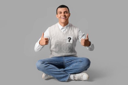 Photo for Young man with paper question mark showing thumbs-up on grey background - Royalty Free Image