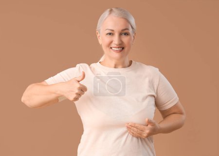 Foto de Mujer madura mostrando el pulgar hacia arriba sobre fondo marrón. Concepto de conciencia sobre el cáncer de mama - Imagen libre de derechos