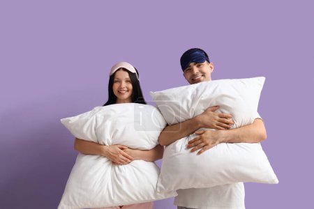 Téléchargez les photos : Jeune couple avec des masques de couchage et des oreillers sur fond lilas - en image libre de droit