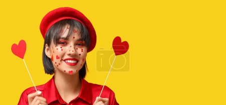 Photo for Stylish young woman with creative makeup for Valentines Day and paper hearts on yellow background with space for text - Royalty Free Image