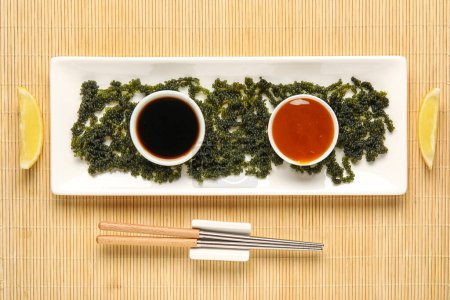 Foto de Composición con plato de algas sanas, salsas y palillos sobre estera de bambú - Imagen libre de derechos