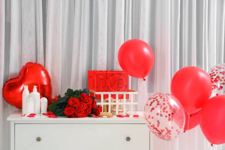 Chest of drawers with bathing accessories, bouquet of roses and heart-shaped balloons in bathroom. Valentine's Day celebration