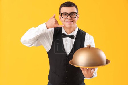 Téléchargez les photos : Male waiter with cloche showing "call me" gesture on yellow background - en image libre de droit