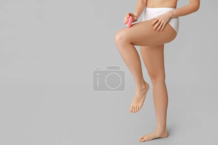 Beautiful young woman epilating her legs with modern epilator on grey background