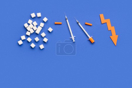 Téléchargez les photos : Cubes de sucre avec seringues pour injection d'insuline et flèche dirigée vers le bas sur fond bleu - en image libre de droit