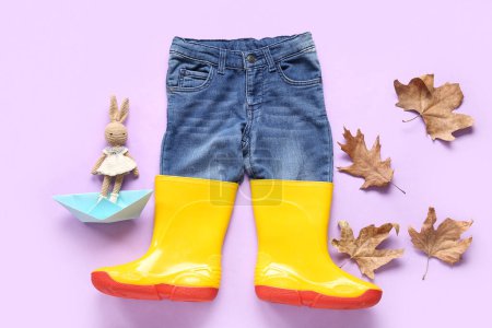 Foto de Botas de goma amarillas con jeans, conejito de juguete y hojas de otoño sobre fondo lila - Imagen libre de derechos