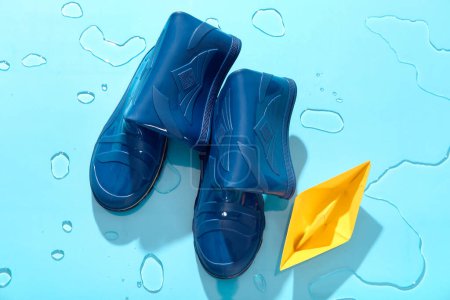 Foto de Elegantes botas de goma azules y barco de papel en agua sobre fondo de color - Imagen libre de derechos