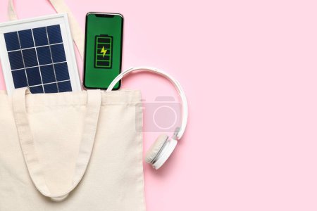 Foto de Bolsa con panel solar portátil, teléfono móvil y auriculares sobre fondo rosa - Imagen libre de derechos