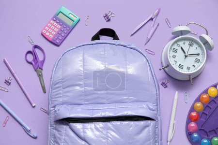 Foto de Mochila escolar con reloj despertador, calculadora y acuarelas sobre fondo lila - Imagen libre de derechos