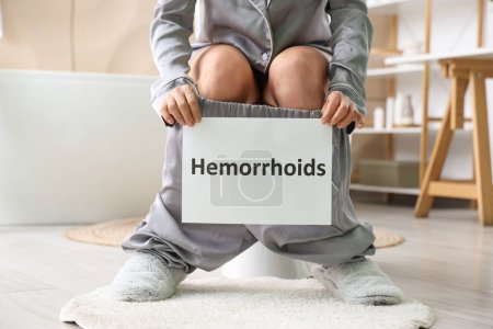 Mujer madura sosteniendo papel con palabra HEMORROIDES en el inodoro en el baño