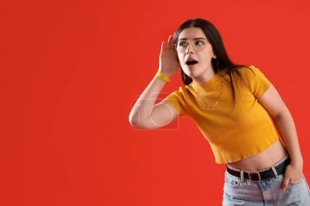 Téléchargez les photos : Jeune femme commérage essayant d'entendre quelque chose sur fond rouge - en image libre de droit