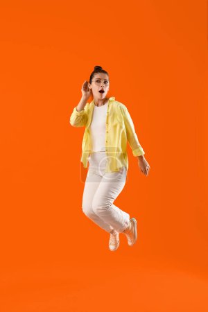 Téléchargez les photos : Jumping jeune femme commérage essayant d'entendre quelque chose sur fond orange - en image libre de droit