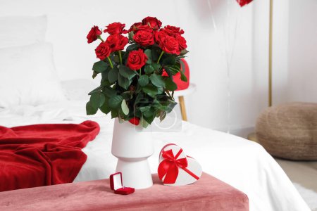 Foto de Ramo de rosas, caja con anillo de compromiso y caja de regalo en forma de corazón en el banco en el dormitorio. Celebraciones del Día de San Valentín - Imagen libre de derechos