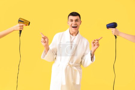 Photo for Shocked young man in bathrobe pointing at female hands with hair dryers on yellow background - Royalty Free Image