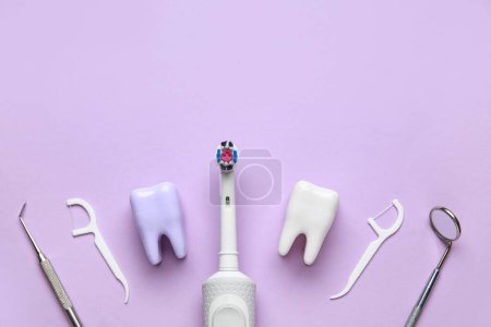 Foto de Modelos de dientes de plástico, cepillo de dientes eléctrico, mondadientes de hilo dental y herramientas dentales sobre fondo lila. Día Mundial del Dentista - Imagen libre de derechos