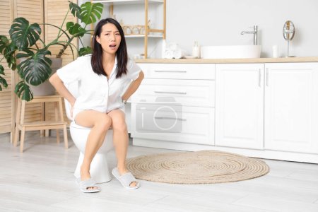 Joven mujer asiática con hemorroides sentado en el inodoro en el baño